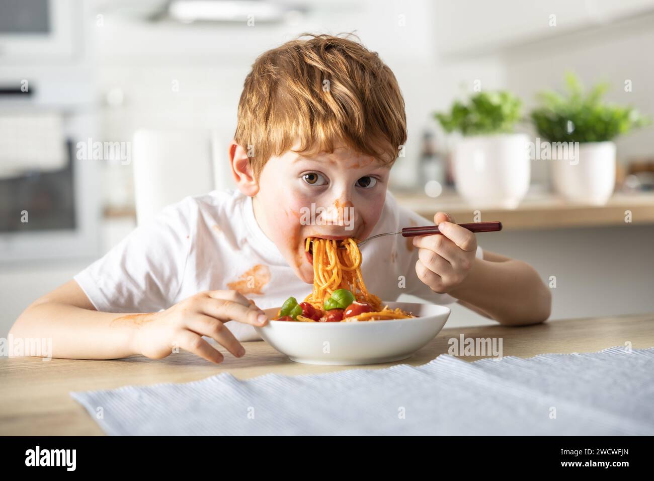 A cute little boy is eating spaghetti bolognese for lunch in the kitchen at home and is covered in ketchup. Stock Photo