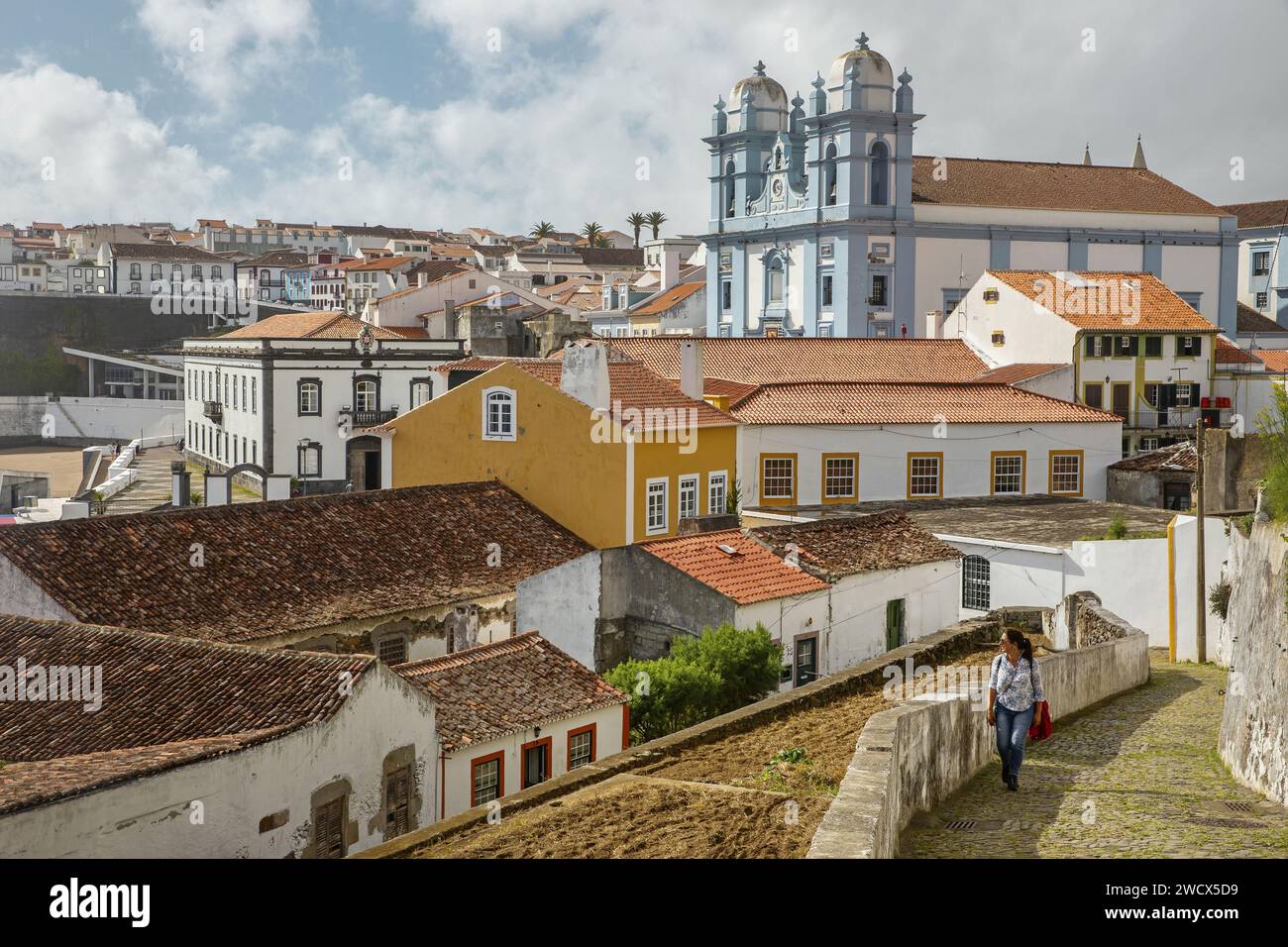 Portugal, Azores archipelago, Terceira island, Angra do Heroismo, woman walking in a cobbled alley in the historic center between colorful colonial buildings with tiled roofs and the Church of Mercy painted in azure blue Stock Photo