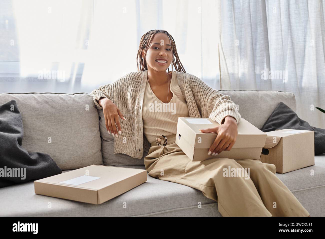 joyful young african american woman in cozy attire sitting on sofa with ...