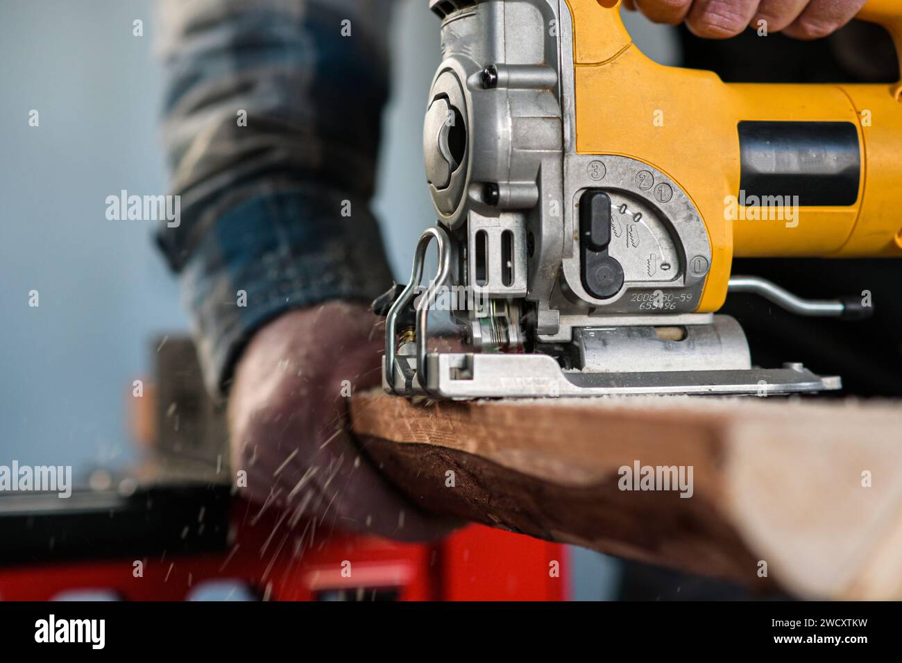 worker in overalls and wearing yellow gloves sawing wood with a reciprocating electric wood saw - close-up view and blurred background. Stock Photo