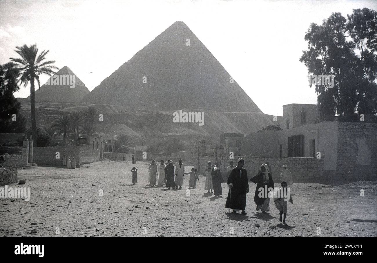 1950s, historical, Egypt, local people walking in a dusty area at Giza, with a pyramid in the background, a classic symbol of this ancient country situated at northeast African coast linking Africa with the Middle East. Its historic monuments include the famous Pyramids at Giza along the fertile Nile river valley. Stock Photo