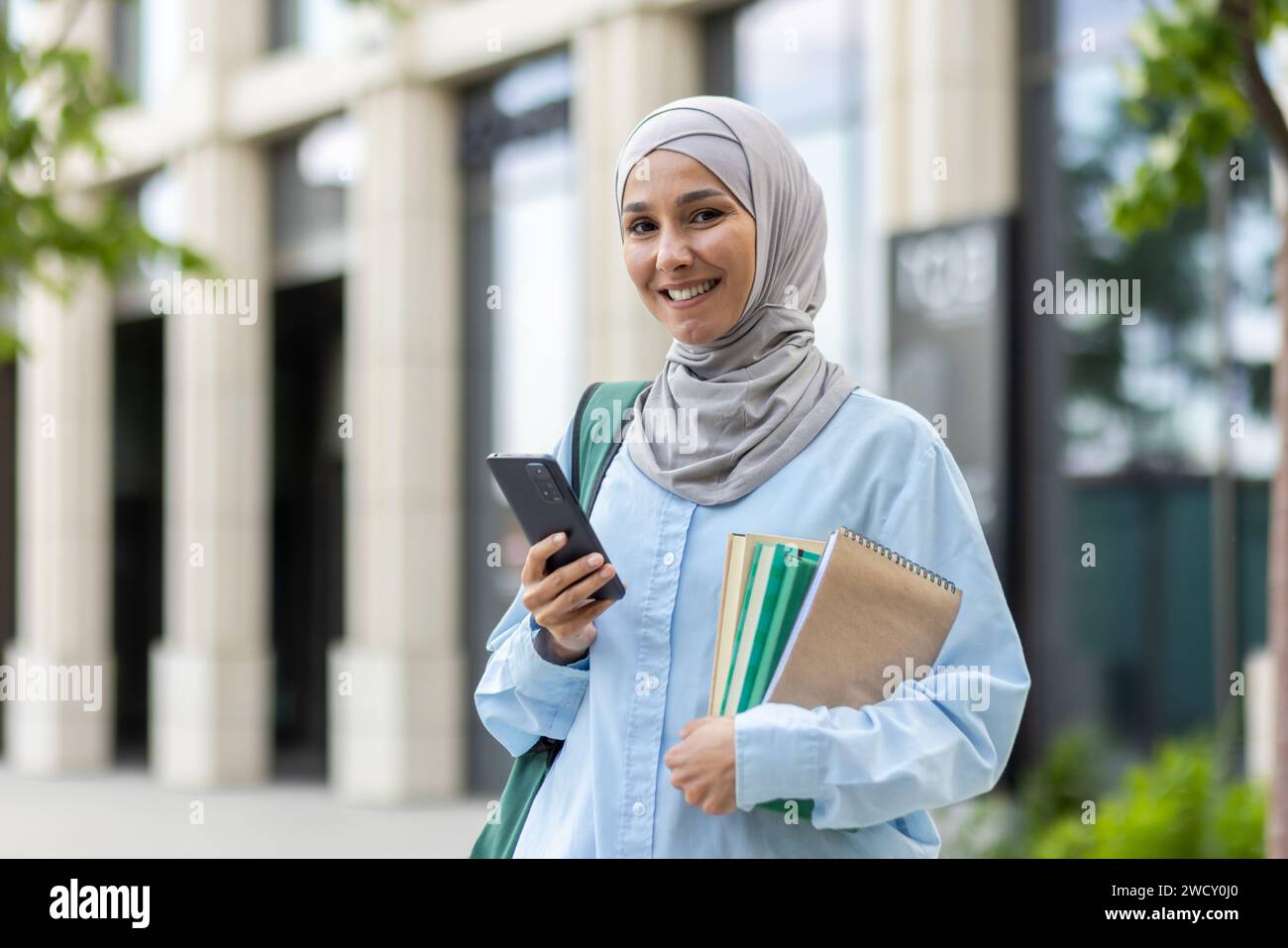 Smiling Arab woman in hijab holding a smartphone and notebook outside ...