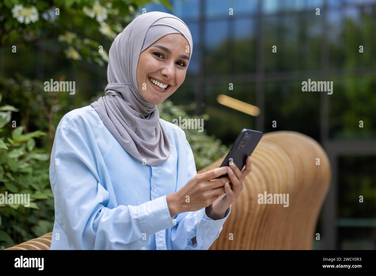 A smiling Muslim woman in hijab operates a smartphone outdoors with an ...