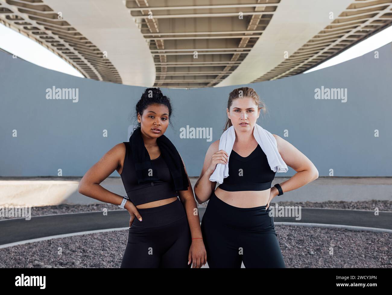 Portrait of two women in black fitness wear with towels on their necks. Young sportswomen are relaxing outdoors. Stock Photo