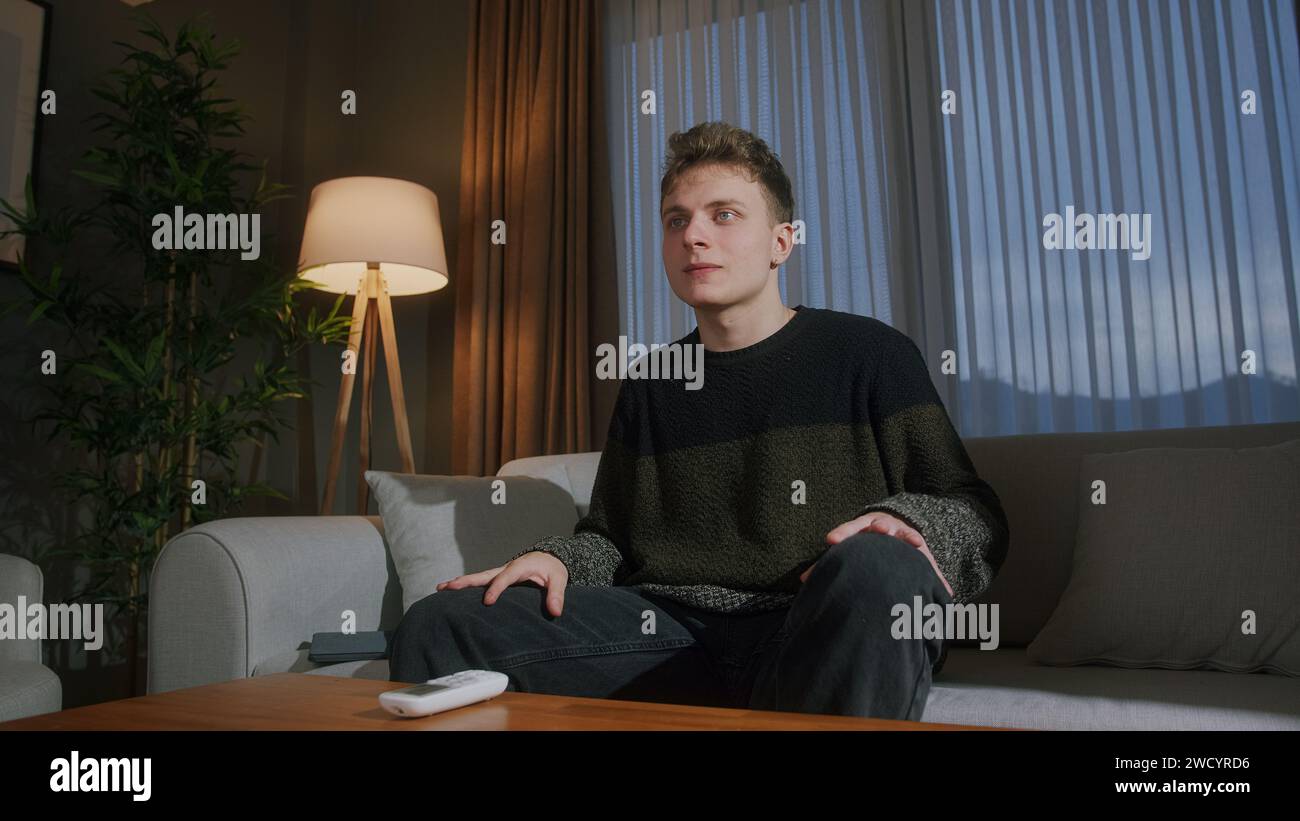 Blonde young man sits on the couch at evening at home, watching a movie on TV Stock Photo