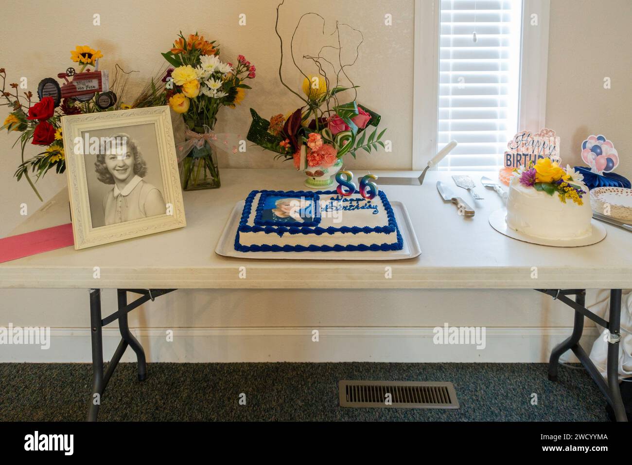 A serving table with two birthday cakes, a photograph, vases of flowers & a Happy Birthday greeting.   USA.b Stock Photo