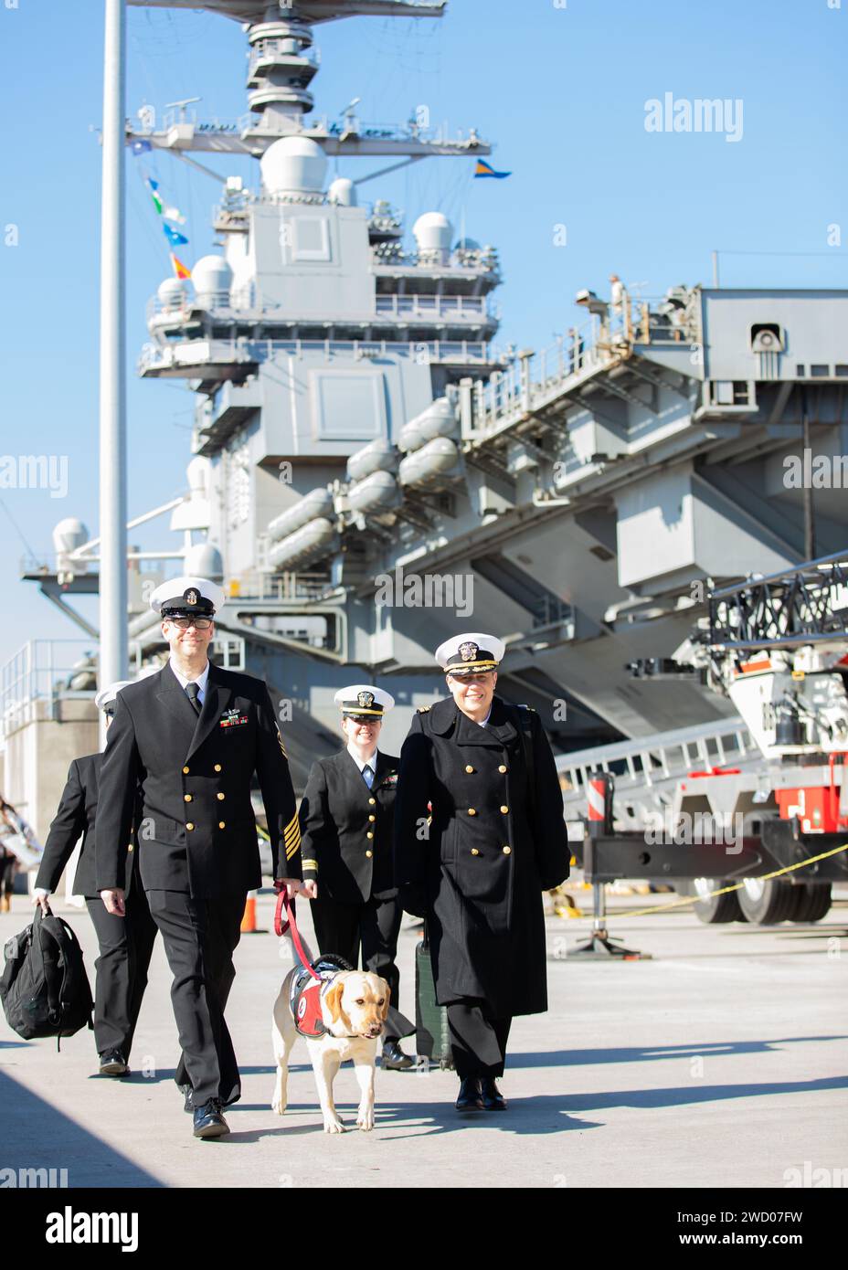 Chief Musician Eric Snitzer, left, assigned to the training department of the world's largest aircraft carrier USS Gerald R. Ford (CVN 78), and Cmdr. Genevieve Clark, assigned Gerald R. FordÕs command religious ministries department, walk the pier with Sage, a four-year-old female Labrador Retriever, deployed aboard Gerald R. Ford through Mutts with a Mission, after the shipÕs eight-month maiden deployment, Jan. 17, 2024. The Gerald R. Ford Carrier Strike Group (GRFCSG) completed a scheduled deployment in U.S. Naval Forces Europe-Africa/U.S. Sixth Fleet area of operations, demonstrating the co Stock Photo