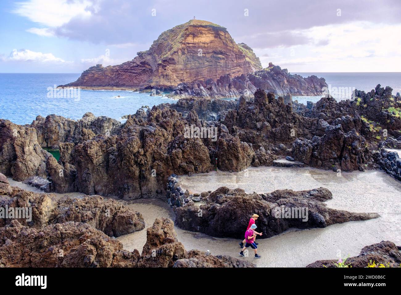 Rocky seascape, two tourists hiking by the ocean at Porto Moniz north coast with natural lava formations, Madeira Island coastline, Portugal Stock Photo