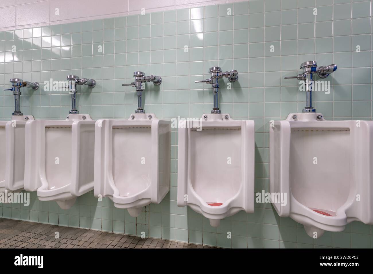 Row of multiple white porcelain ceramic urinals in an older men's bathroom with green tile. Stock Photo