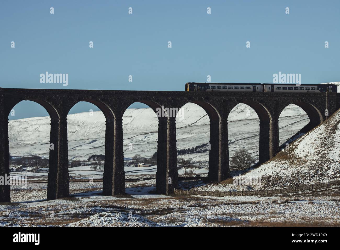 A Northern train crossing the railway on the Ribblehead Viaduct in the snow in the Yorkshire Dales National Park. Taken on a beautiful blue sky day. Stock Photo