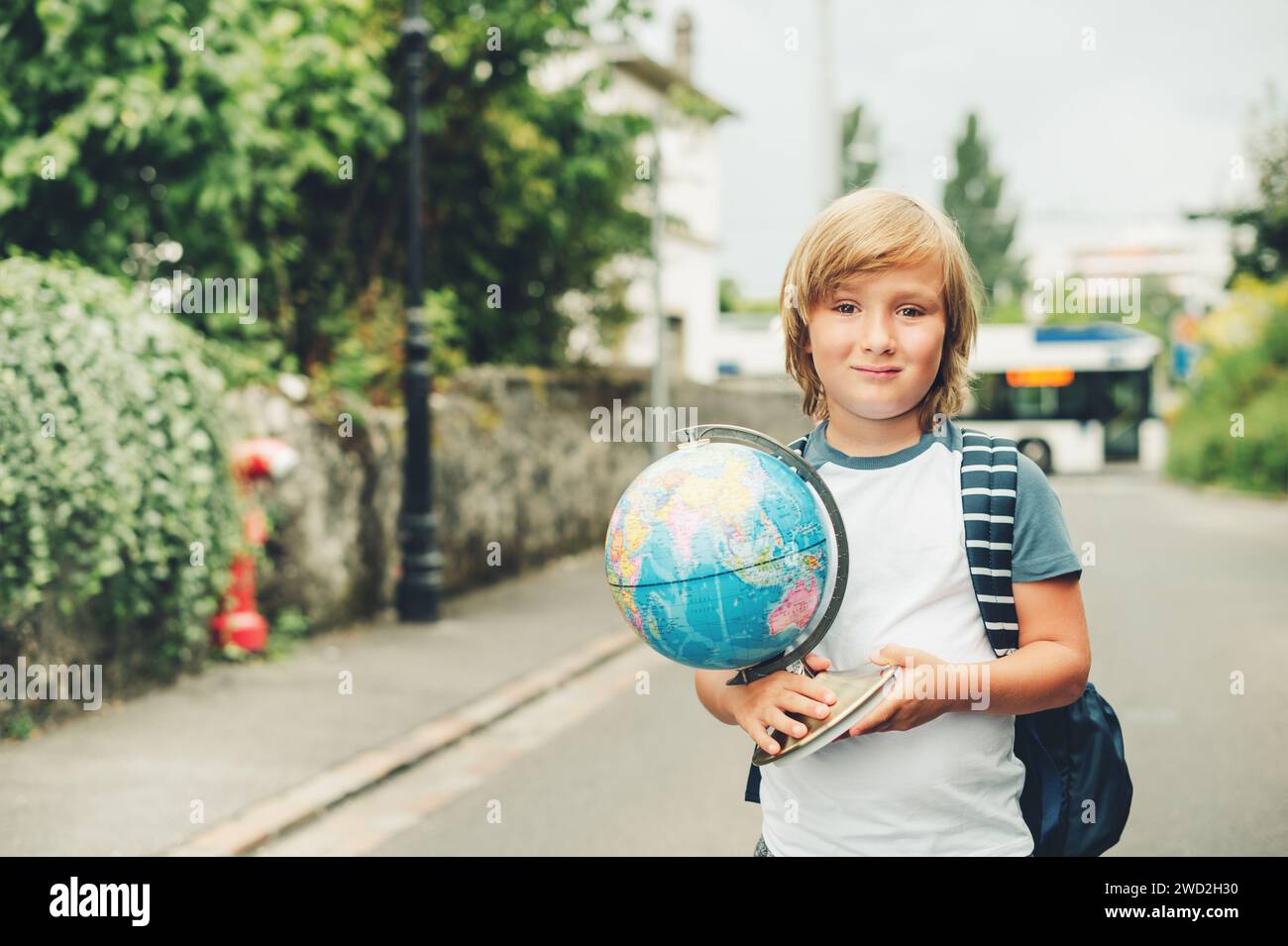 Outdoor portrait of funny little kid boy wearing backpack and holding world globe. Back to school concept. Film look filter image Stock Photo
