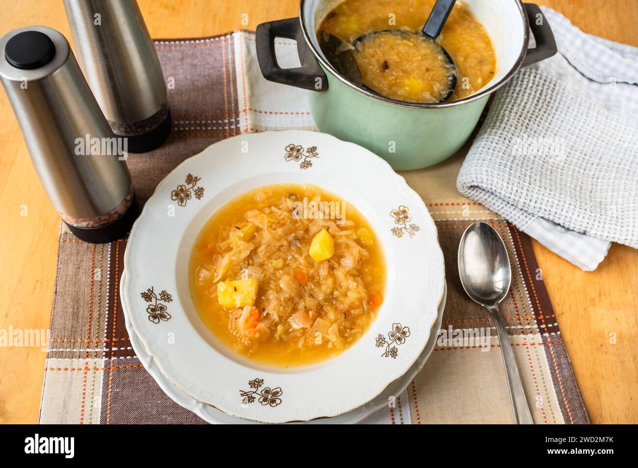 Cabbage soup with potato in decorative plate, pot with soup and ladle, 2 spice rack, towel on table. Stock Photo