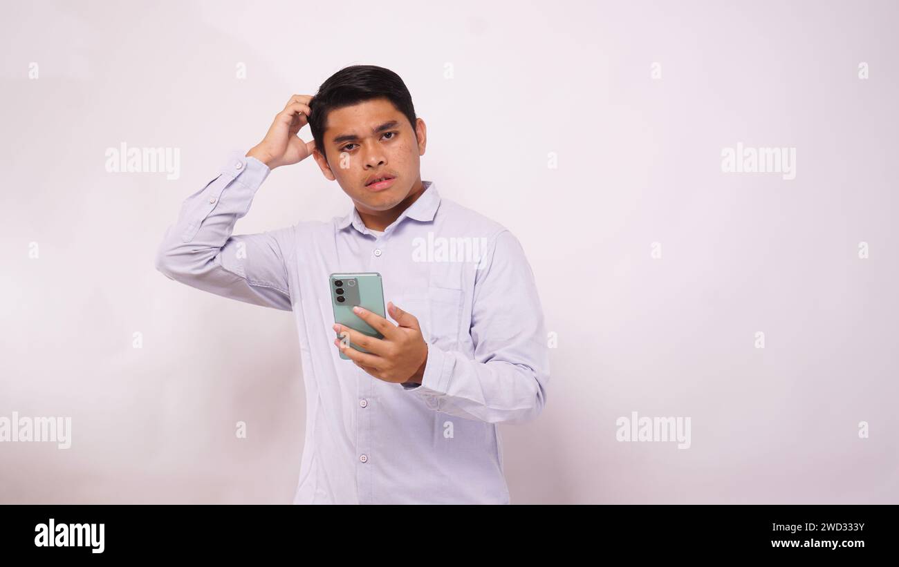 Asian man scratch his head while holding mobile phone and showing confused face expression against white background Stock Photo