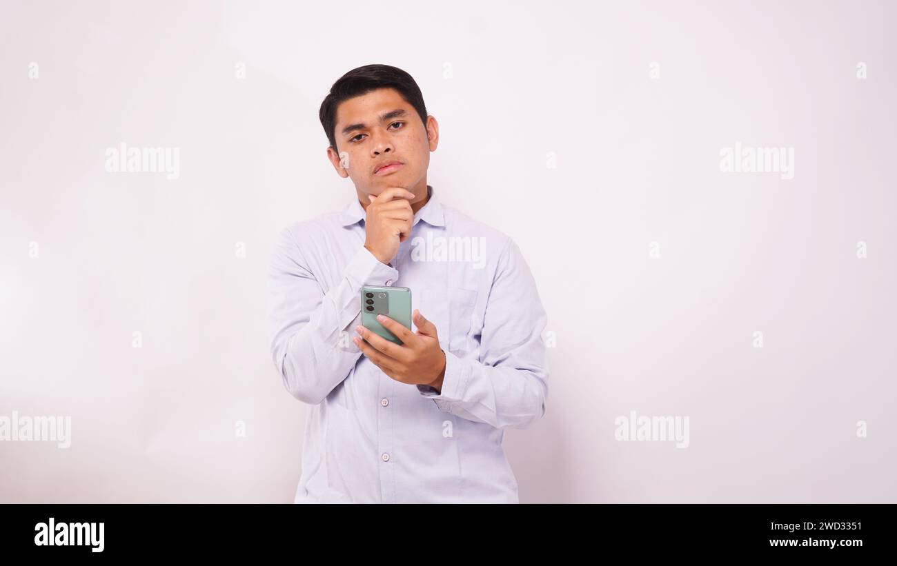 Asian man scratch his head while holding mobile phone and showing confused face expression against white background Stock Photo