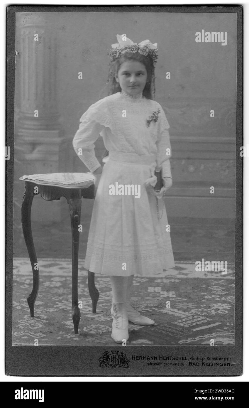 Studio portrait of a teenage girl in a white dress with a book in her left hand, standing near the table. Photo studio Herman Hentschel. Bad Kissingen Stock Photo