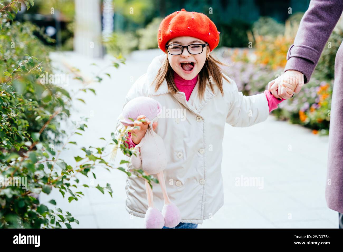 Happy laughing little girl in eyeglasses with special needs holding mother's hand on a walk in autumn time outdoor. Stock Photo