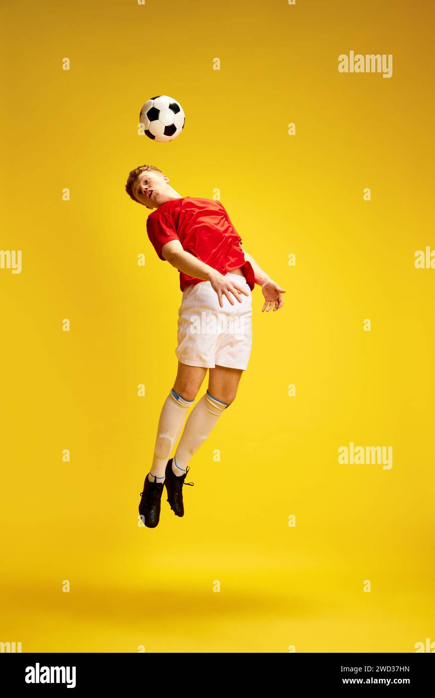Full-length image of young man in uniform, soccer player, enthusiast in motion, training, hitting ball with head against yellow studio background Stock Photo