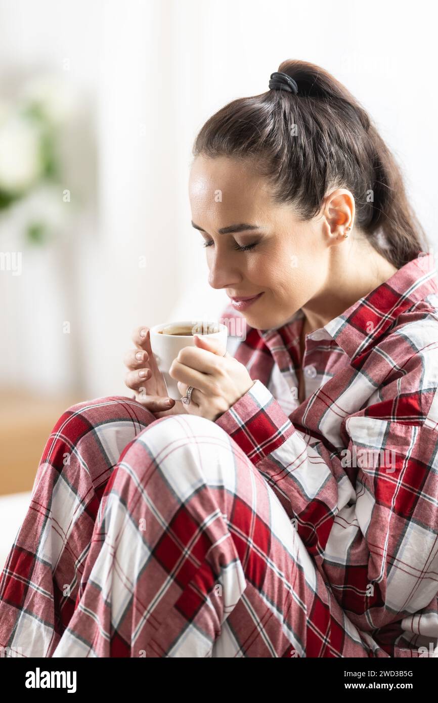 Young woman in red checkered pajama sitting on sofa and drinking coffee at the morning in livng room. Stock Photo