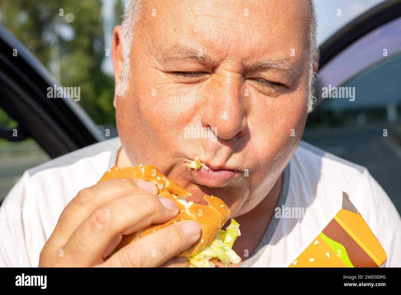 Grown man enjoying burger by the side of a car. The concept of fast food and takeaway food. Big juicy burger with cheese from McDonalds in the hand. Kiev, Ukraine, July 08 2023 Copyright: xDmytroxNikolaienkox Stock Photo