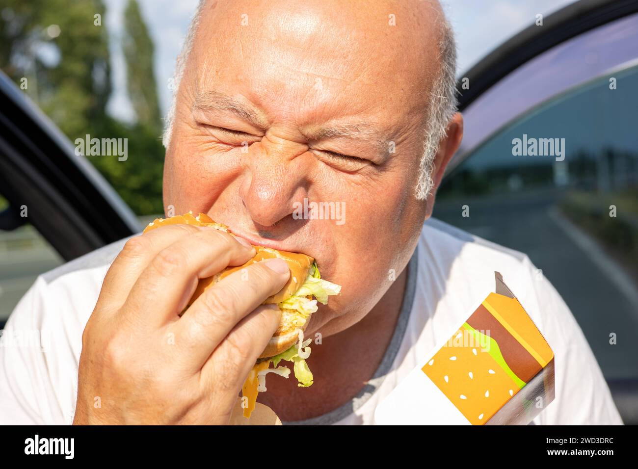 Grown man enjoying burger by the side of a car. The concept of fast food and takeaway food. Big juicy burger with cheese from McDonalds in the hand. Kiev, Ukraine, July 08 2023 Copyright: xDmytroxNikolaienkox Stock Photo