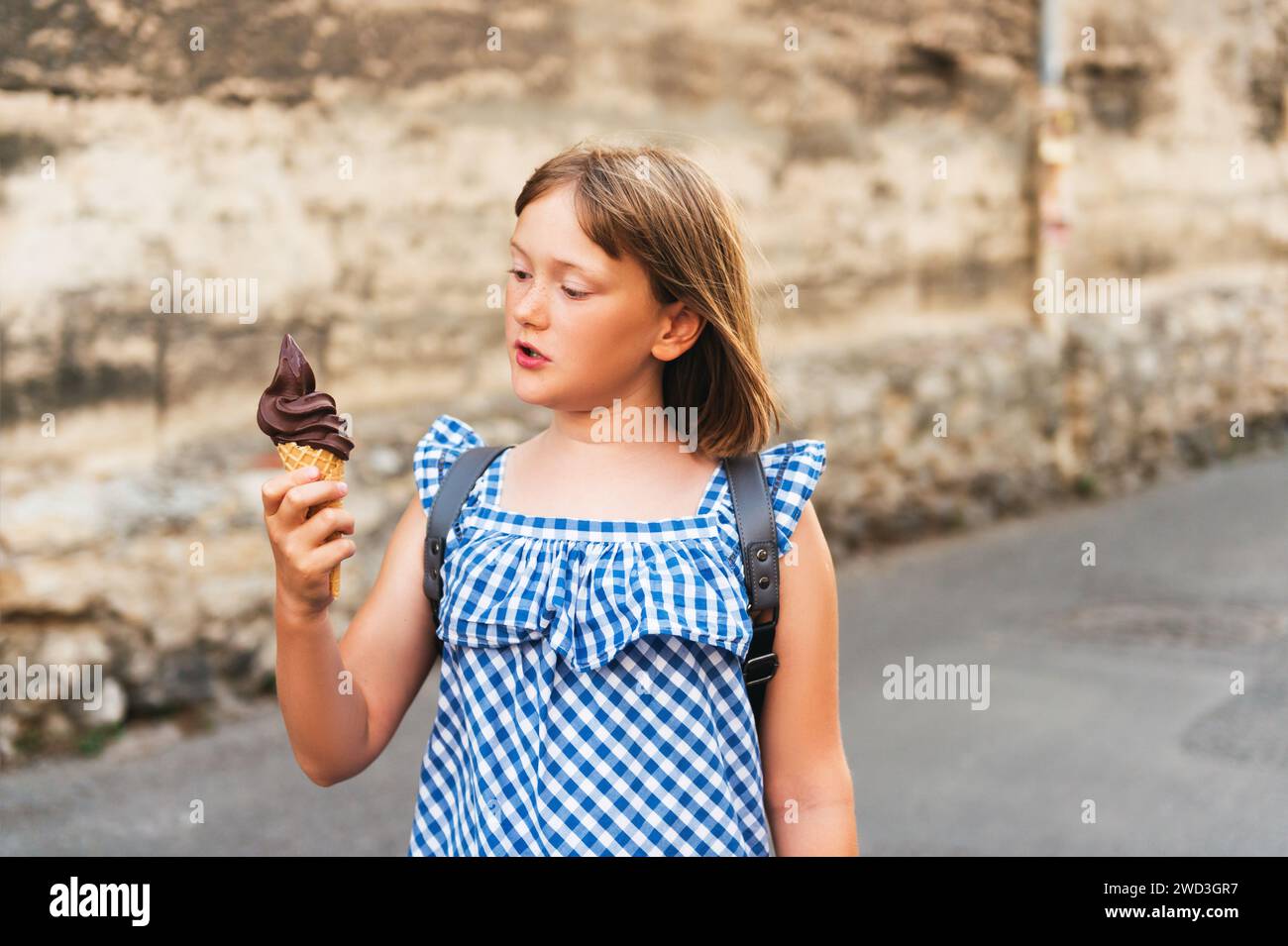 Cute little girl eating chocolate ice cream outdoors, wearing blue gingham dress and backpack, travel with kids Stock Photo