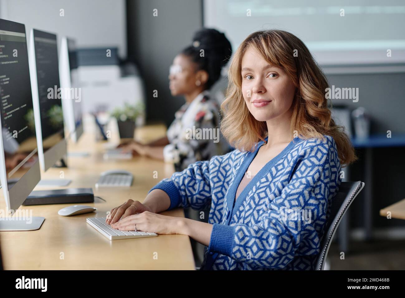 Caucasian female coder sitting at her desk working with data on computer and smiling at camera Stock Photo
