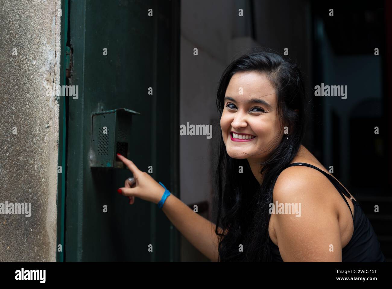 Portrait of beautiful young woman with black hair ringing the doorbell of a house and smiling. Person traveling. Stock Photo