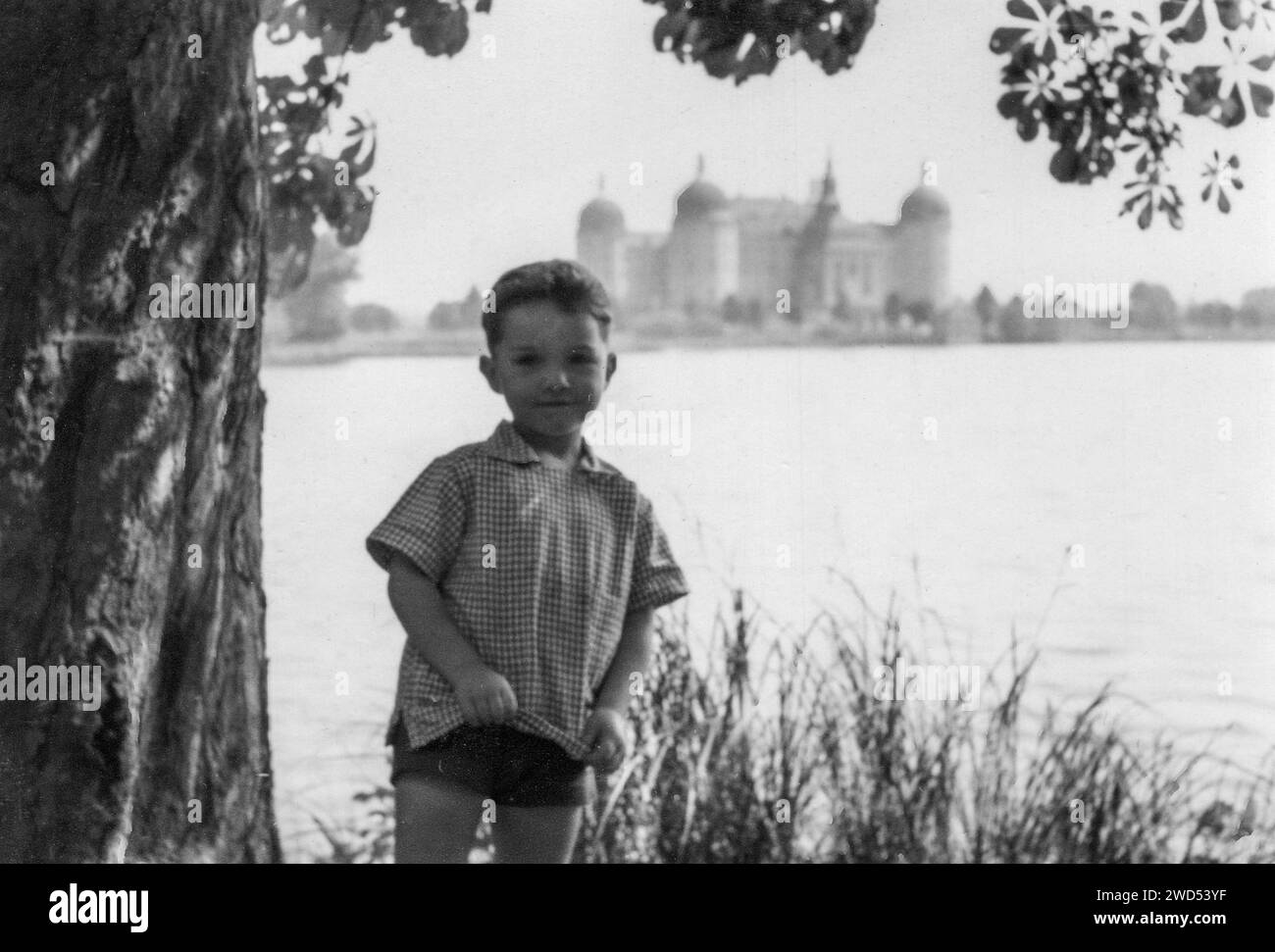 An antique photo with shows a small boy in shorts and a short-sleeved shirt posing against the backdrop of a lake in front of Moritzburg Castle. Dresd Stock Photo