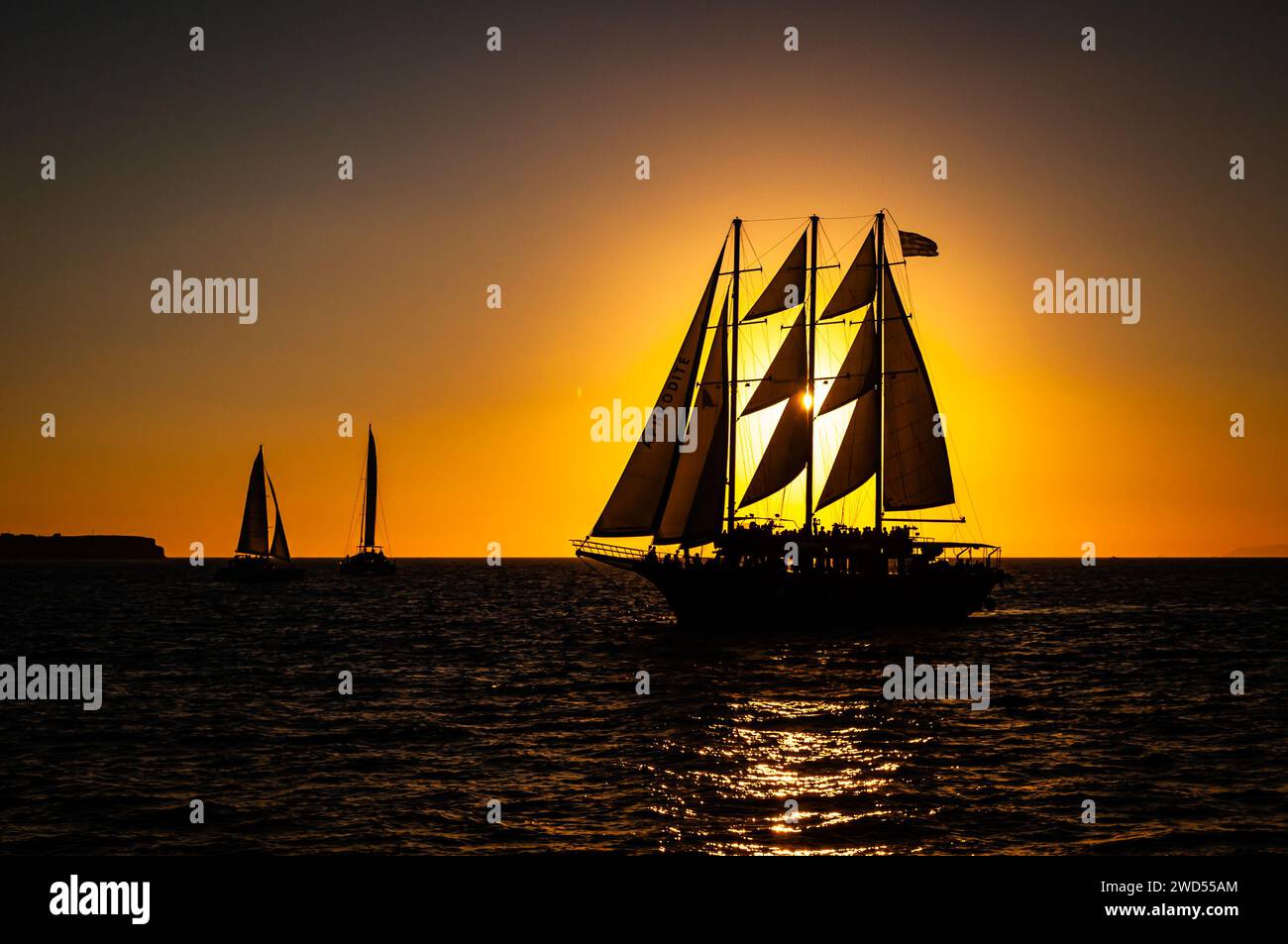 An orange-hued sunset casts a warm glow on the silhouette of several sailboats gliding across the calm surface of the water Stock Photo
