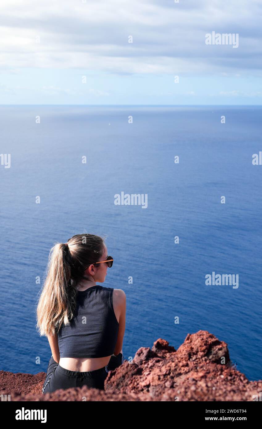 In the awe-inspiring landscape of Red Mountain in El Medano, Tenerife, a woman finds solace and connection as she perches atop a striking red rock ove Stock Photo