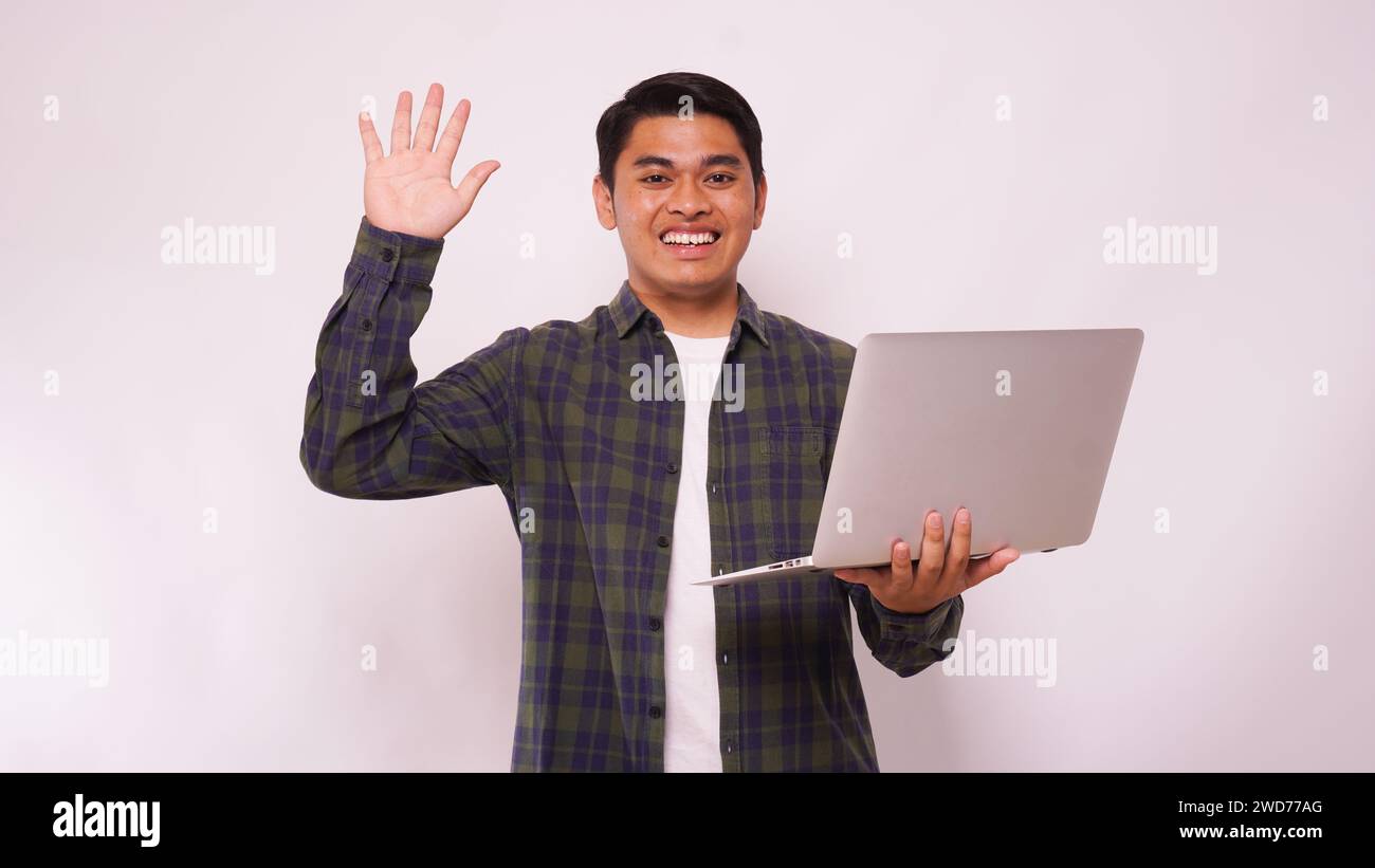 Asian student waving hand while holding laptop against white background Stock Photo