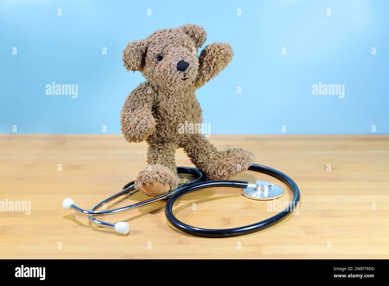 Little brown teddy bear standing on a stethoscope and waving, health care for children, pediatric medicine concept, blue background with copy space, s Stock Photo