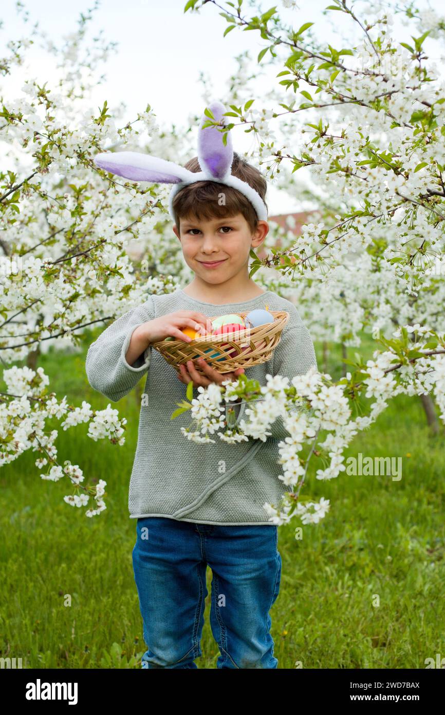 Easter egg hunt. Cute preschool boy wearing bunny ears holding full basket of Easter eggs in hands at Easter egg hunt in garden. Portrait against back Stock Photo