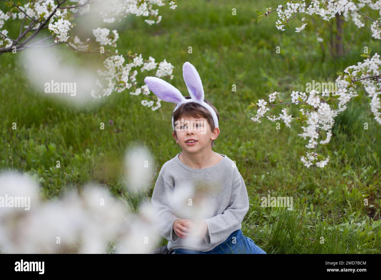 Cute preschool boy with bunny ears in garden. smiling child siting on green lawn against background of trees blooming with white flowers. Happy easter Stock Photo