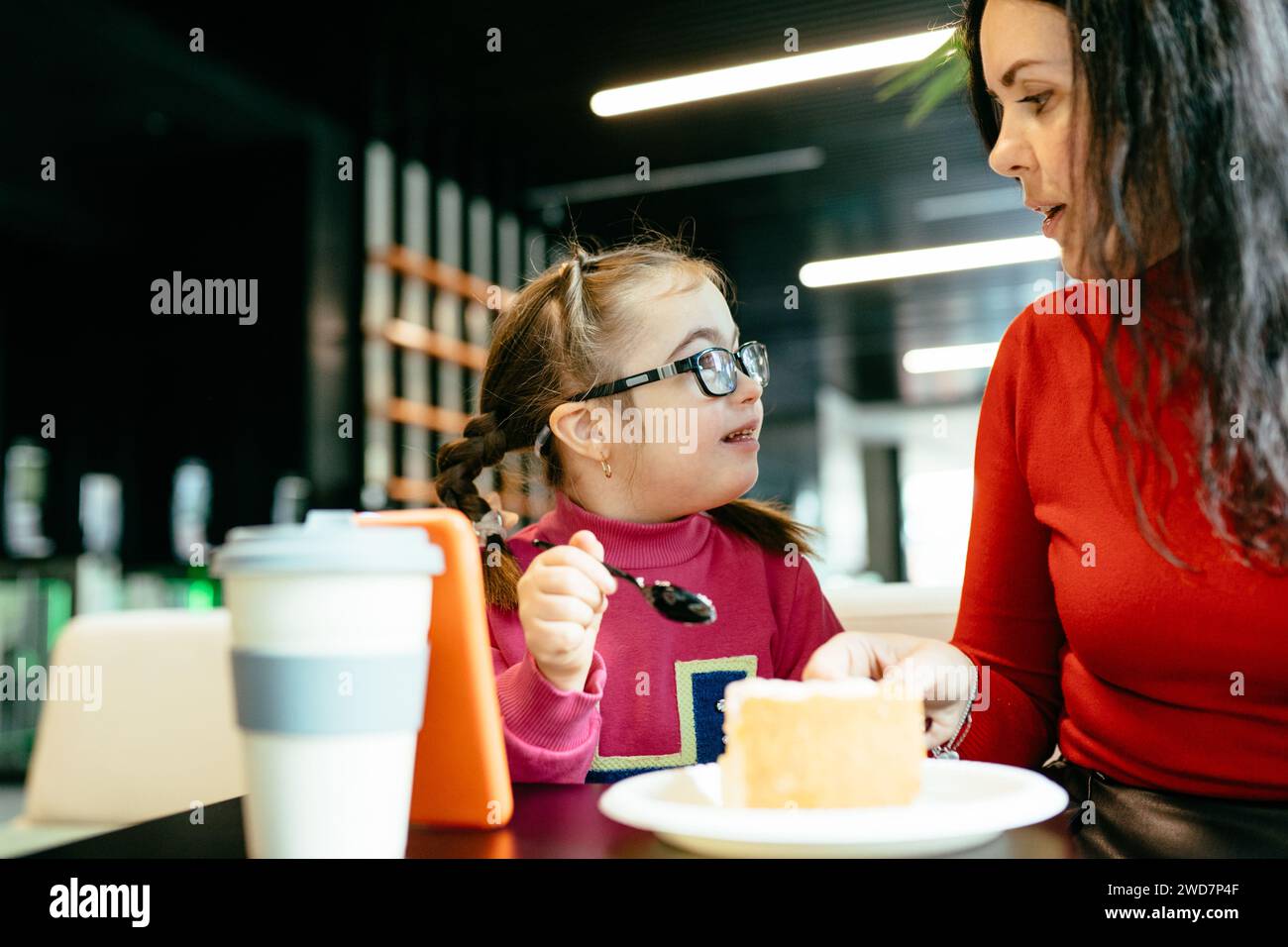 Portrait of happy mother smiling to each other cute girl with down syndrome eating cake in cafe. Happy family moments concept. Stock Photo