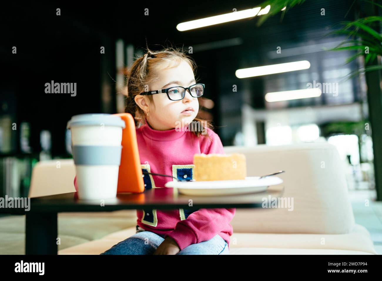Cute little girl in eyeglasses using smartphone eating cake at cafe. Stock Photo