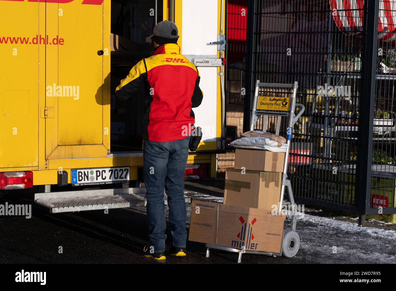 Lieferdienst in Aktion: DHL-Zusteller bereitet Paketlieferung bei REWE vor. , Deutschland, Rheinland-Pfalz, Speyer, 19.01.2024, Auf dem Foto ist ein DHL-Zusteller zu sehen, der neben einem offenen, gelben Lieferwagen steht und Pakete auf einem Transportwagen vor sich hat. Im Hintergrund ist ein REWE-Schild erkennbar, was darauf hinweist, dass die Lieferung in der Nähe eines REWE-Supermarktes stattfindet. *** Delivery service in action DHL delivery driver prepares parcel delivery at REWE, Germany, Rhineland-Palatinate, Speyer, 19 01 2024, The photo shows a DHL delivery driver standing next to a Stock Photo