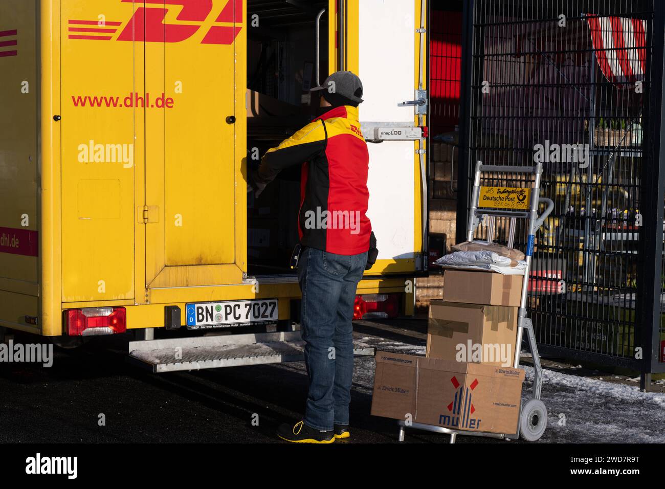 Lieferdienst in Aktion: DHL-Zusteller bereitet Paketlieferung bei REWE vor. , Deutschland, Rheinland-Pfalz, Speyer, 19.01.2024, Auf dem Foto ist ein DHL-Zusteller zu sehen, der neben einem offenen, gelben Lieferwagen steht und Pakete auf einem Transportwagen vor sich hat. Im Hintergrund ist ein REWE-Schild erkennbar, was darauf hinweist, dass die Lieferung in der Nähe eines REWE-Supermarktes stattfindet. *** Delivery service in action DHL delivery driver prepares parcel delivery at REWE, Germany, Rhineland-Palatinate, Speyer, 19 01 2024, The photo shows a DHL delivery driver standing next to a Stock Photo