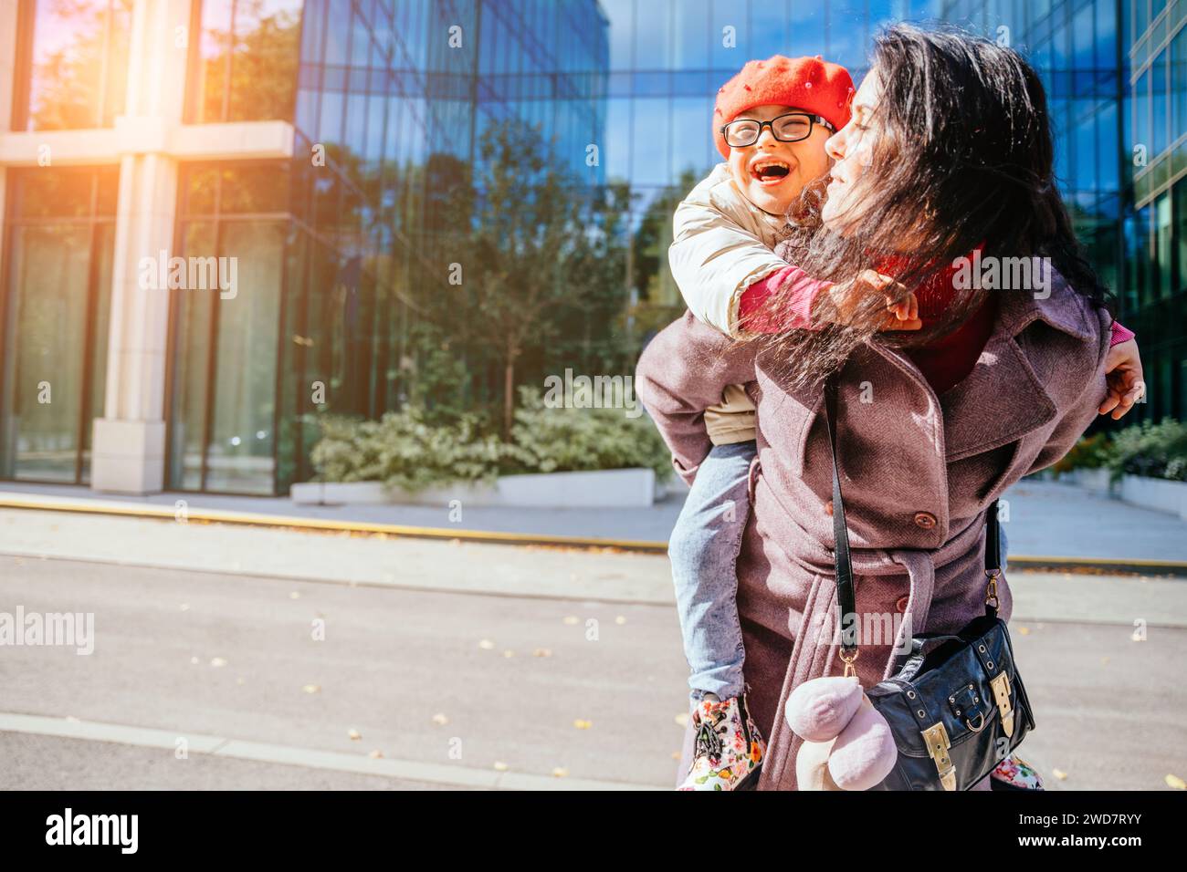 Cute laughing daughter on a piggy back ride with her brunette happy mother in autumn time with modern building at city street on background. Stock Photo