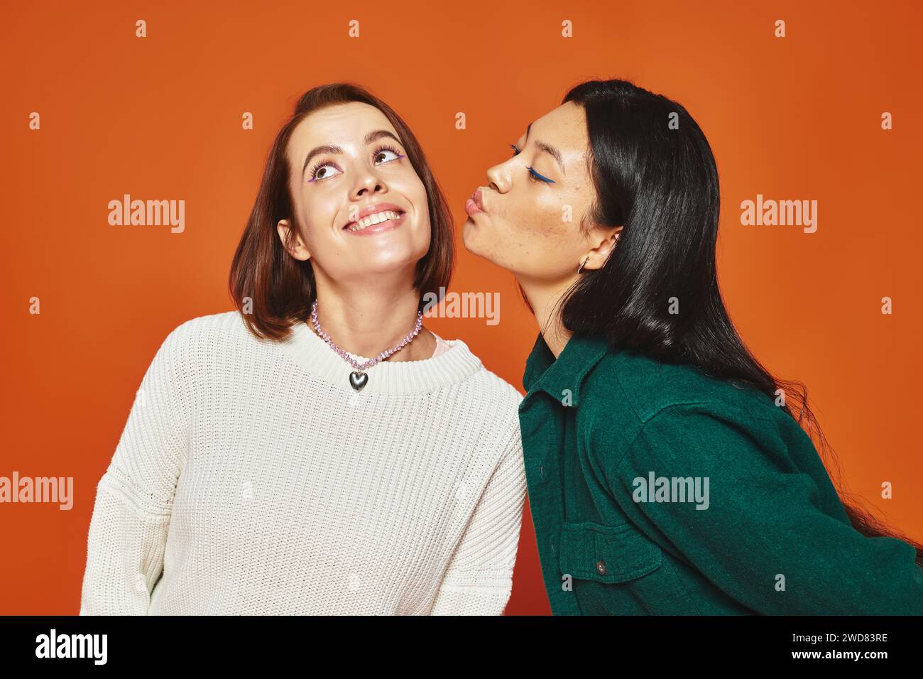 cute asian woman kissing cheek of her cheerful female friend on orange ...