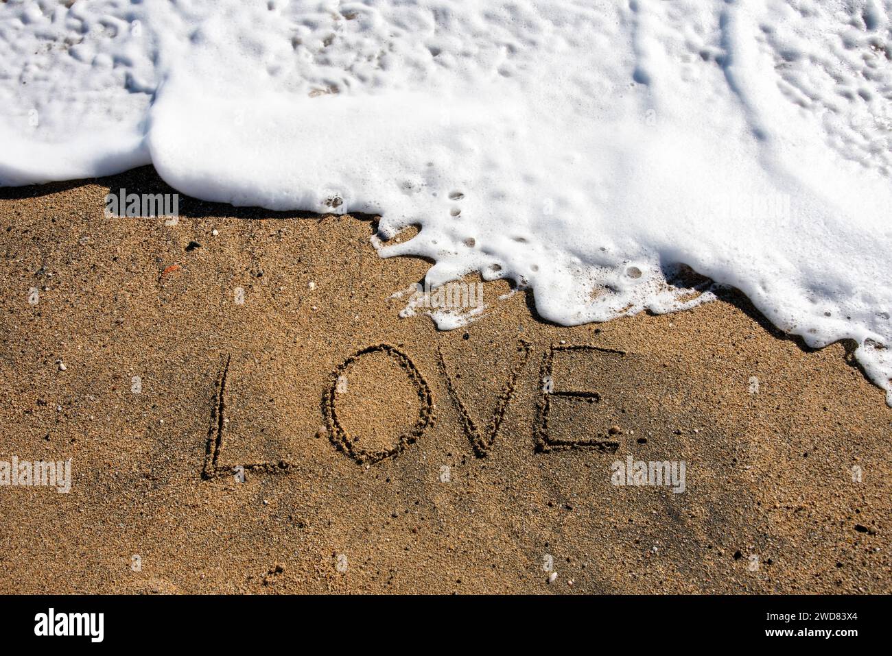 Love unveiled: 'Amor' etched in beach sands, embraced by a gentle wave, a poetic moment along the shore Stock Photo