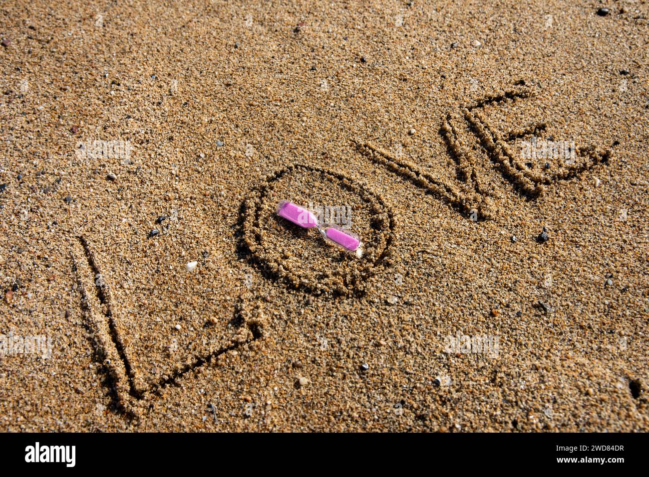 Timeless love: 'Love' inscribed on the beach, cradling a pink-hued hourglass, where romance meets the sands of eternal moments. Stock Photo