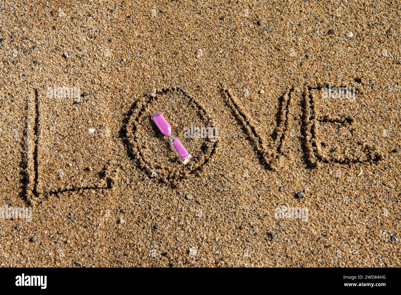Timeless love: 'Love' inscribed on the beach, cradling a pink-hued hourglass, where romance meets the sands of eternal moments Stock Photo