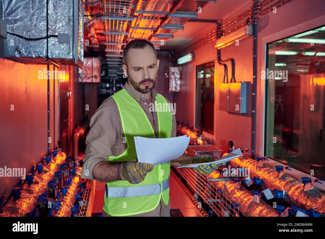 concentrated hardworking man in safety clothes looking attentively at his paperwork, data center Stock Photo