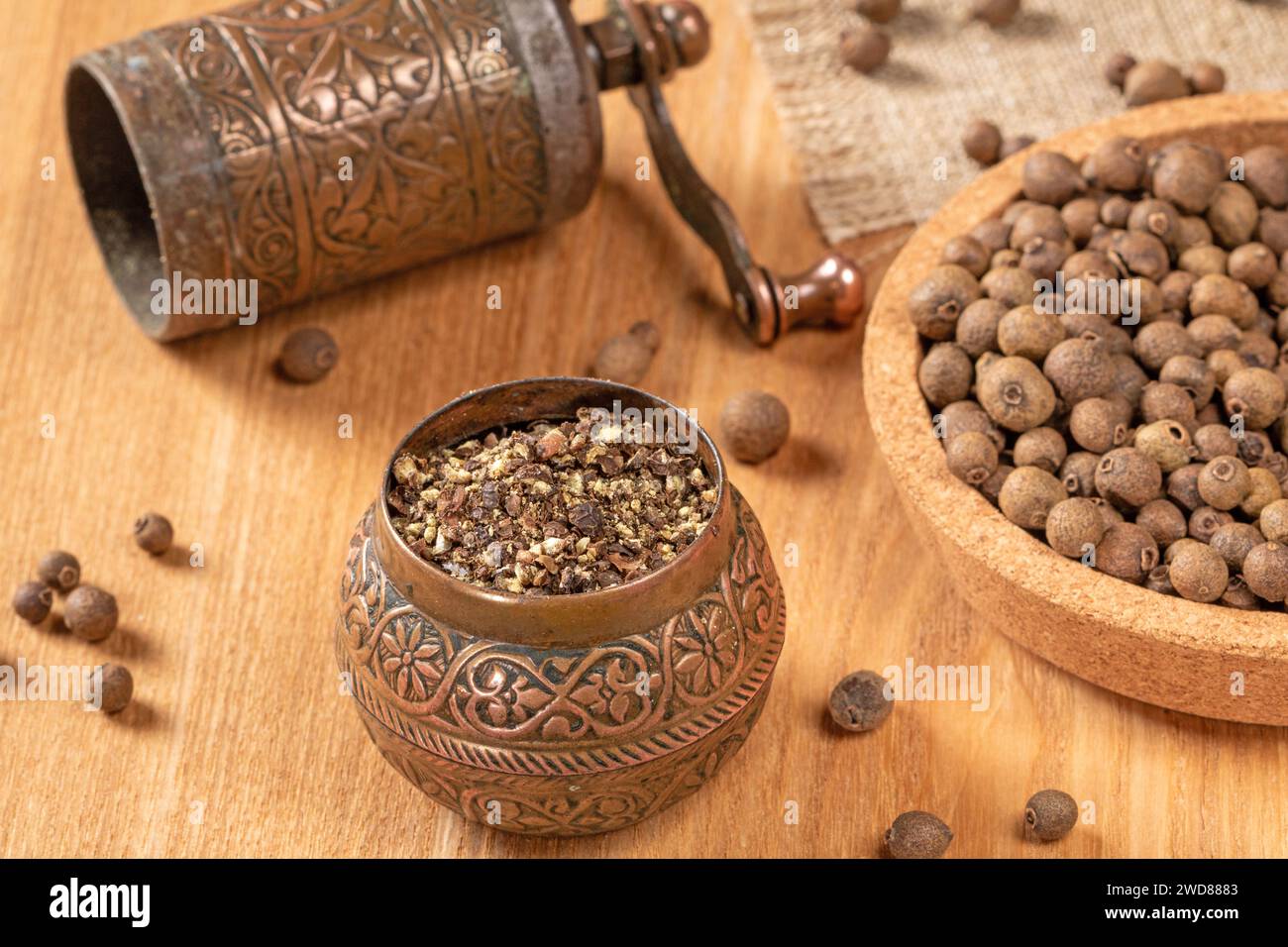 A close-up of a traditional black pepper mill filled with whole peppercorns, resting on a textured wooden background. The warm lighting enhances the r Stock Photo