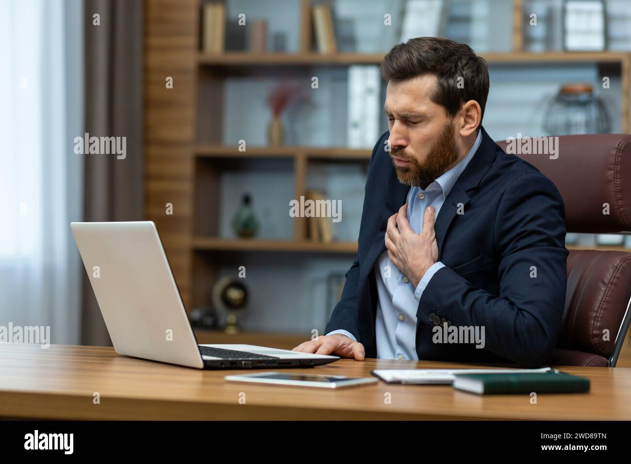 Mature businessman in a home office setup looks distressed, clutching his chest as if experiencing discomfort or pain. Stock Photo