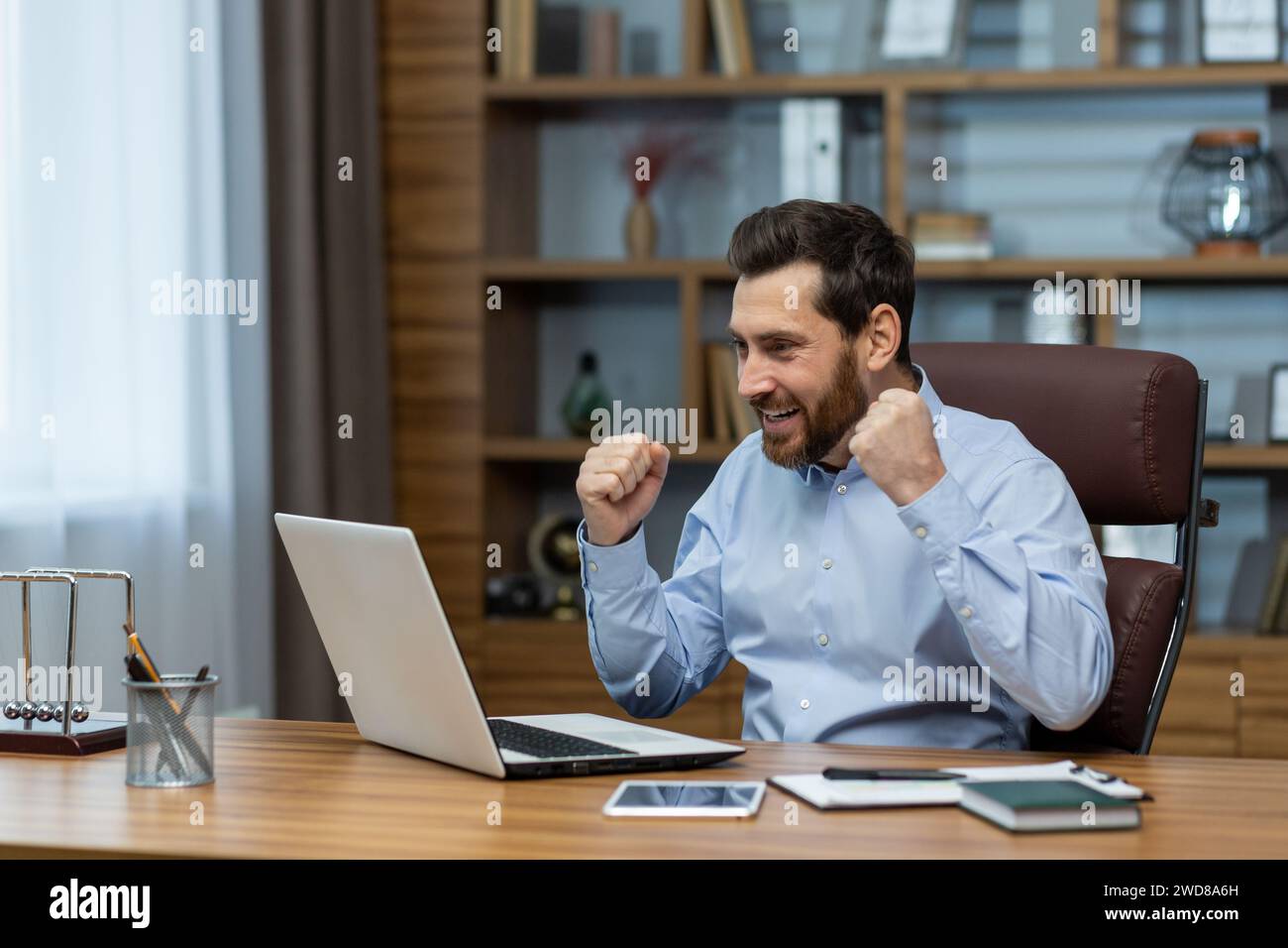 Exuberant mature businessman with a beard showing excitement and success at his home office desk, laptop screen visible. Stock Photo
