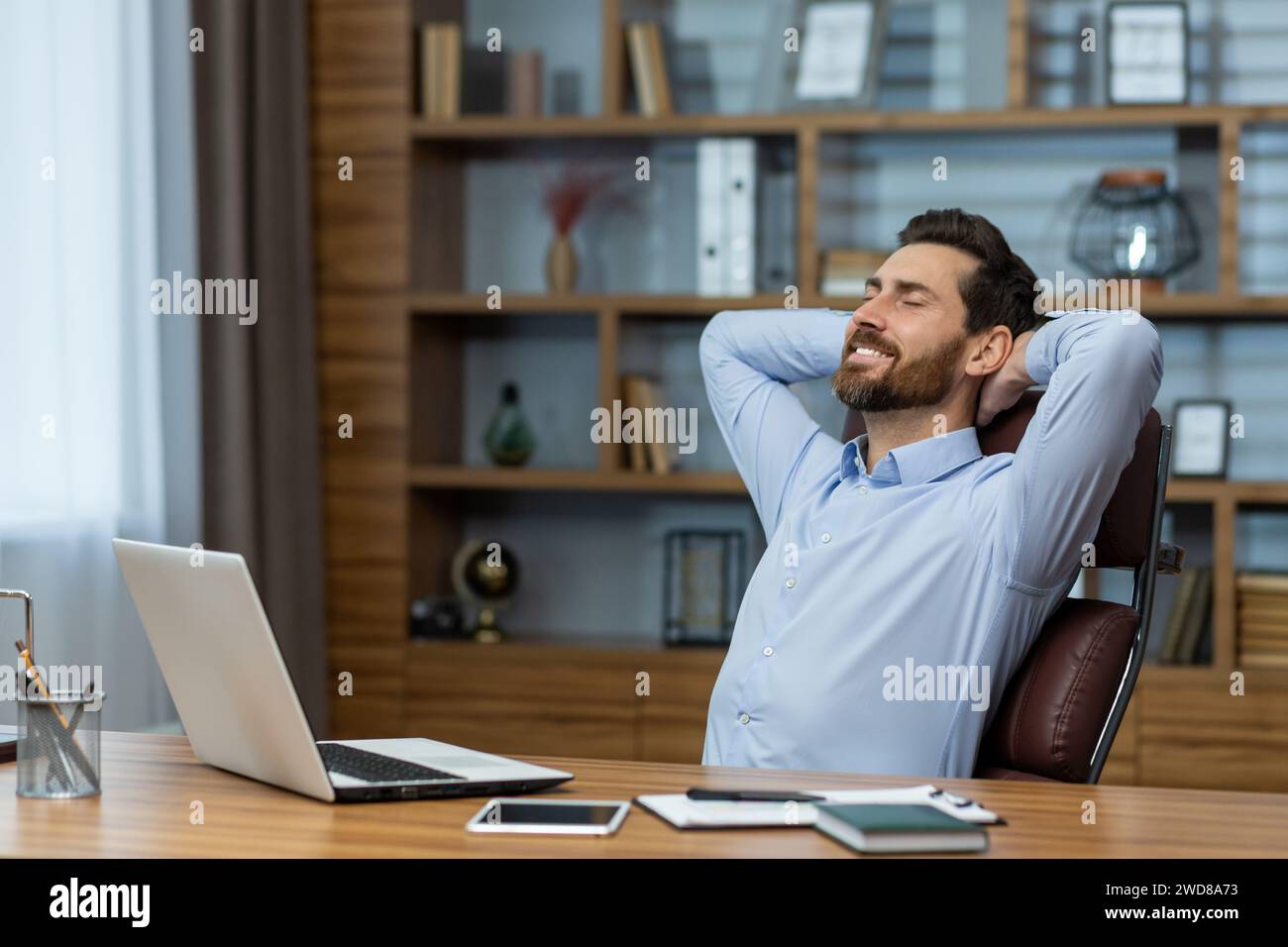 Content mature businessman taking a break, relaxing with hands behind his head in a well-organized home office setting. Stock Photo