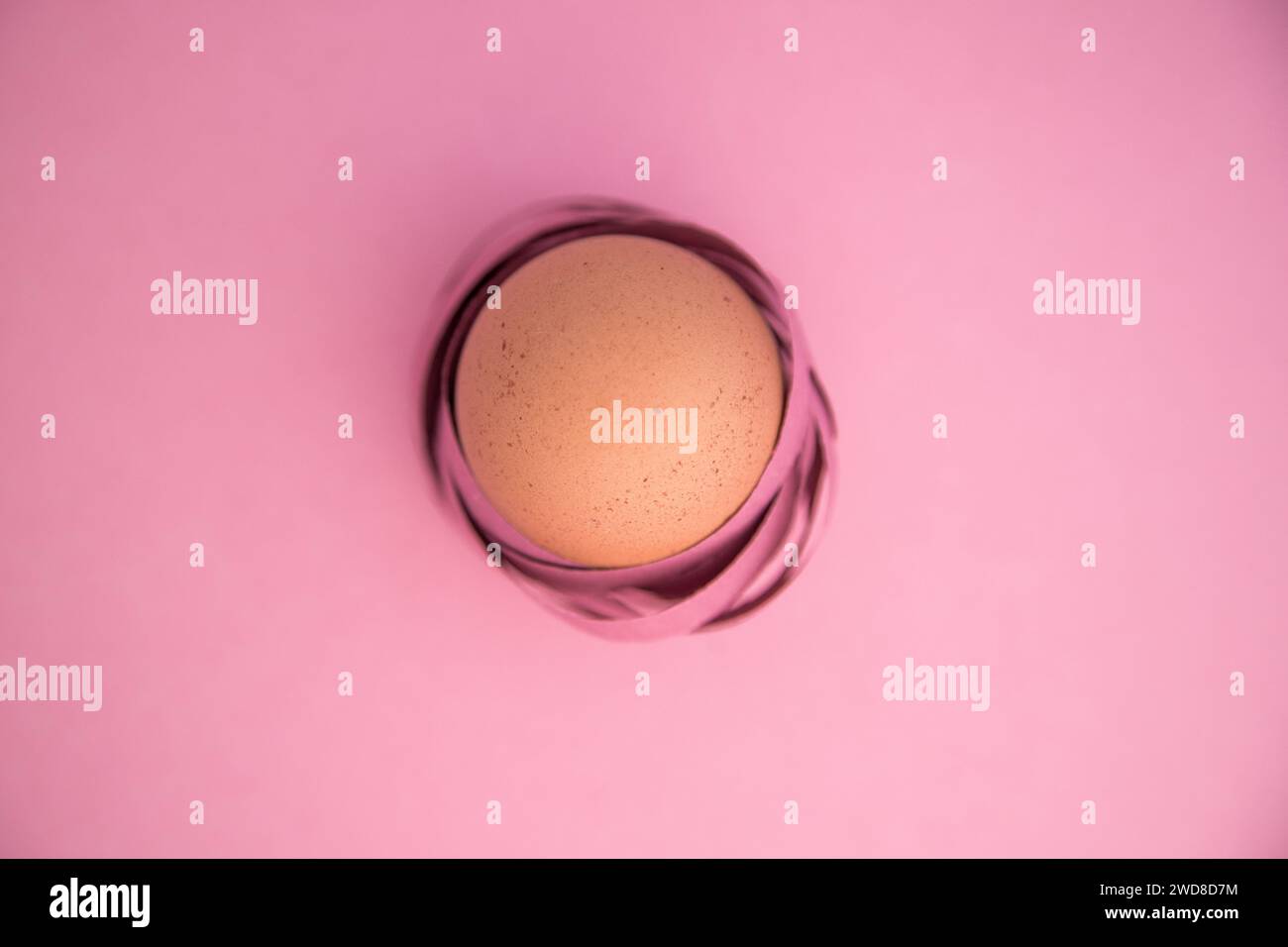 Happy Easter. Minimalist close-up of an egg wrapped in colored ribbons as if it were a nest. Isolated on pink background. Stock Photo