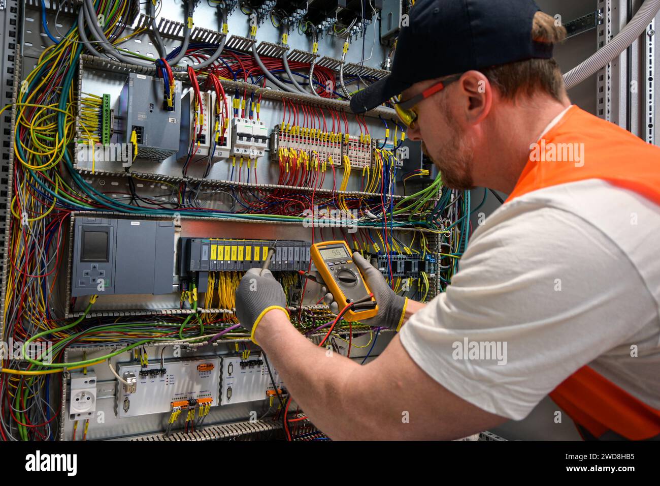 an electrician in a cap, yellow glasses and an orange vest measures electric current with a digital multimeter on a distribution box. Stock Photo